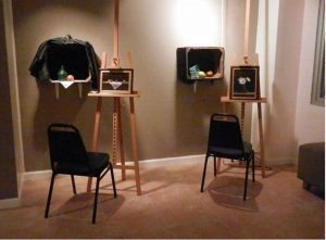 Festőállványok, mellettük, posztamenseken álló, “árnyék dobozok” a beállított csendéletekkel 