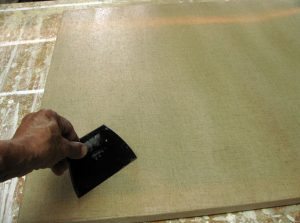 Ezt a lépést, csak akkor kell elvégeznünk, ha vásznat impregnálunk, enyvvel, vagy PVA ragasztóval. Egy életlen késsel, vigyázva, hogy a vásznat ne sértsük meg, ne vágjuk ki, “Lehúzzuk” a felesleges enyvet a vászonról, a kés éle kb. 45 fokos szögben van a vászonra fektetve, és közben erősen bele is “passzírozzuk” az enyvet a vászon szövet apró kis lyukacskáiba. Ezzel a technikával amellett, hogy egyenletesebb lesz az enyv bevonata a vásznon, egyszersmind, tömítjük ezeket a kis lyukakat, így amikor az alapozót felhordjuk, az szebben, egyenletesebben fog elterülni a vásznon. 